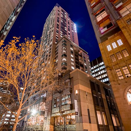 Amora Hotel Jamison Сідней Екстер'єр фото The photo shows a tall building illuminated at night, surrounded by other structures in an urban environment. The foreground features a tree with bare branches, suggesting it's possibly autumn or winter. The building itself has a modern design, with 