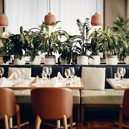 Amora Hotel Jamison Сідней Екстер'єр фото The photo shows an elegant restaurant interior. There are several tables neatly arranged, each set with glasses, white napkins, and light-colored tableware. The seating consists of brown chairs and a bench along the wall. Lush green plants are promin