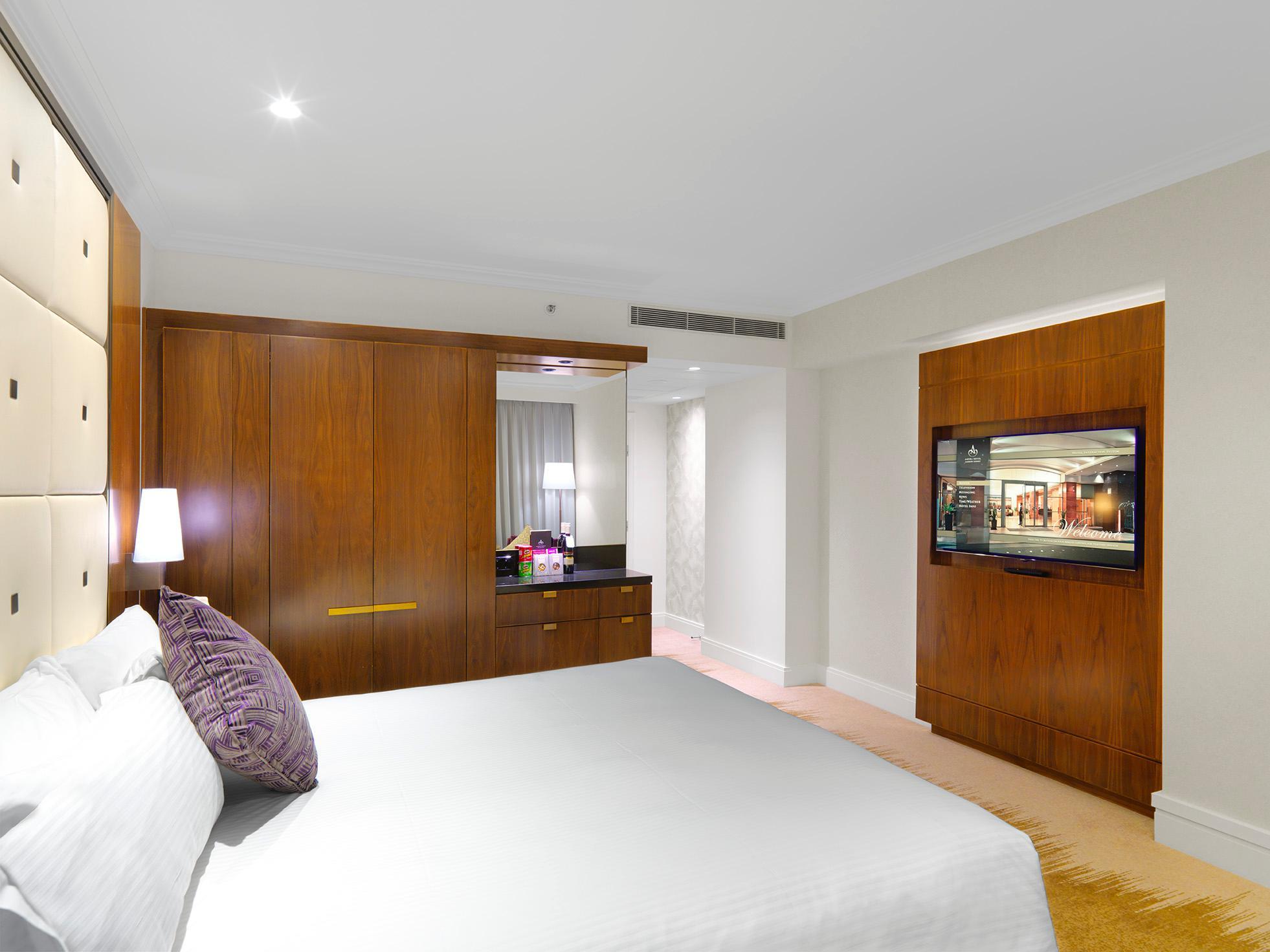 Amora Hotel Jamison Сідней Екстер'єр фото The photo shows a modern hotel room featuring a neatly made bed with white linens and a decorative purple pillow. The room has wooden finishes, including a built-in wardrobe and a chest of drawers. There is a wall-mounted television on one side and s