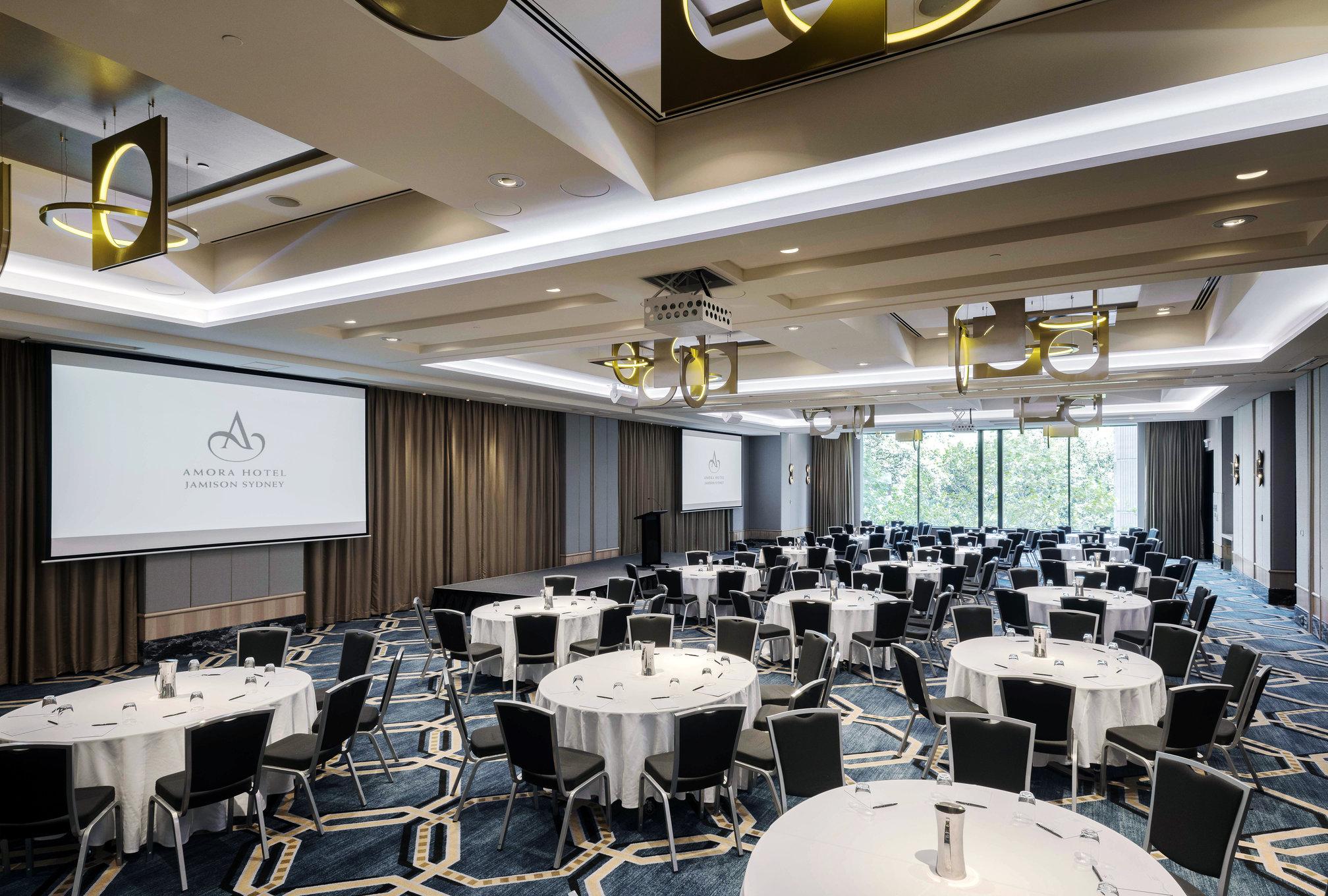 Amora Hotel Jamison Сідней Екстер'єр фото The photo shows a spacious conference or event room. There are multiple round tables arranged with white tablecloths and black chairs around them. The room has a modern design with elegant lighting fixtures hanging from the ceiling. A large screen is