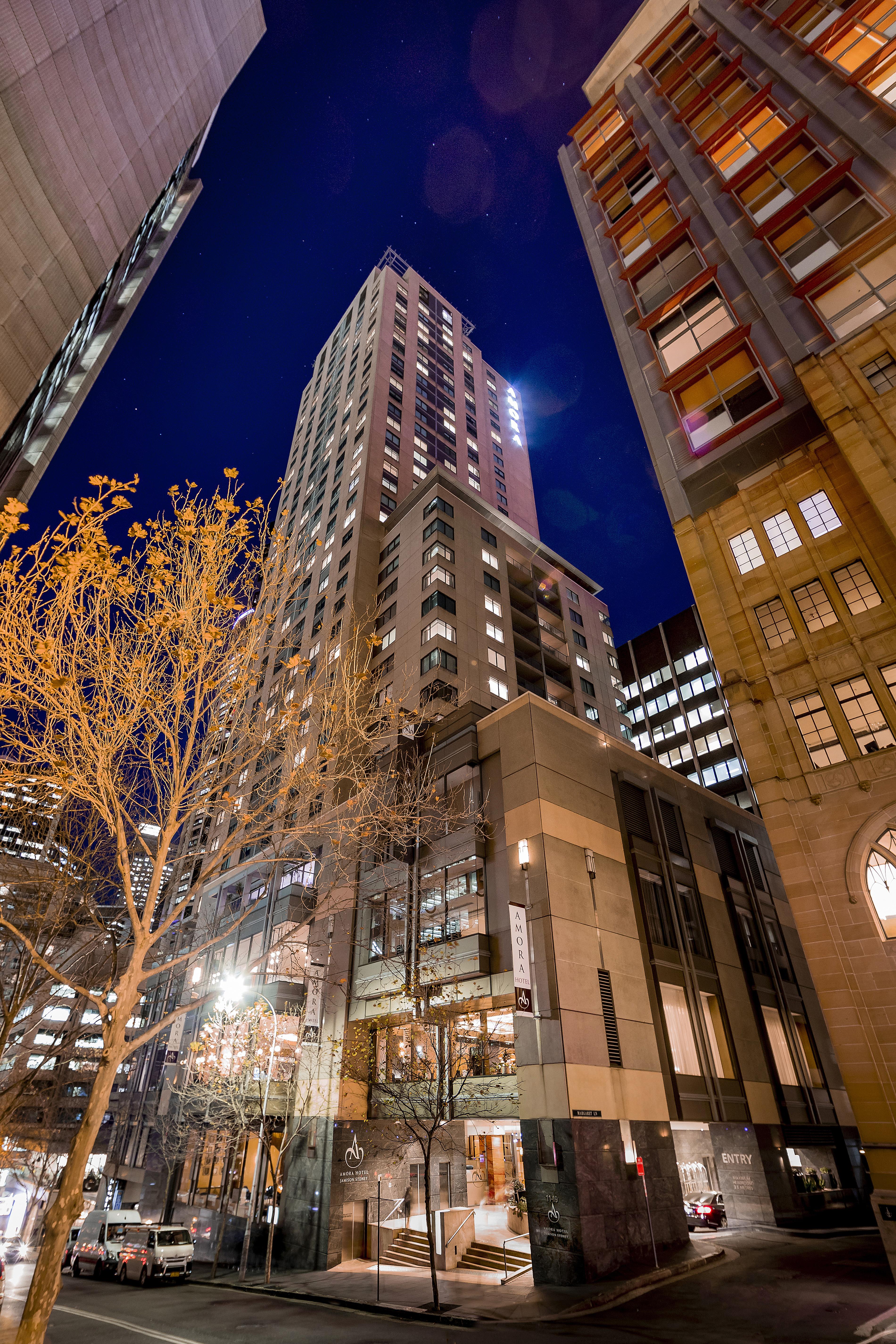 Amora Hotel Jamison Сідней Екстер'єр фото The photo shows a tall building illuminated at night, surrounded by other structures in an urban environment. The foreground features a tree with bare branches, suggesting it's possibly autumn or winter. The building itself has a modern design, with 