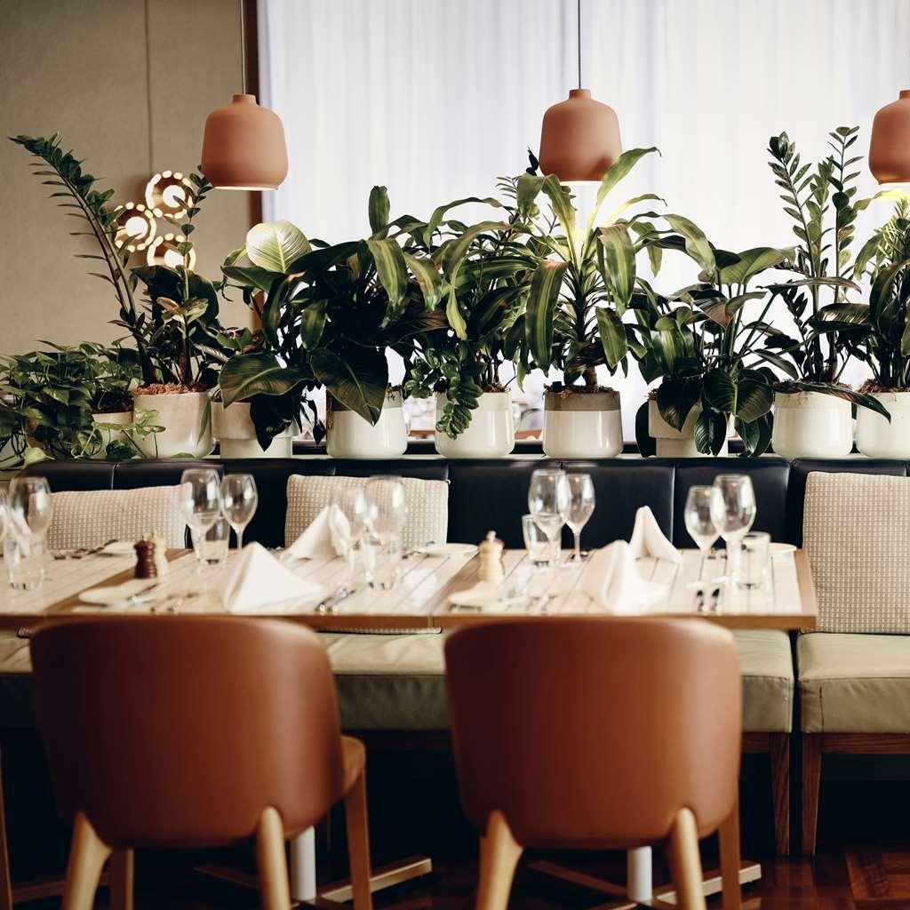 Amora Hotel Jamison Сідней Ресторан фото The photo shows a stylish restaurant interior featuring a warm and inviting atmosphere. There are neatly arranged tables with wooden surfaces, each set with glasses, napkins, and seasonings. Surrounding the tables are large, green plants in white pot