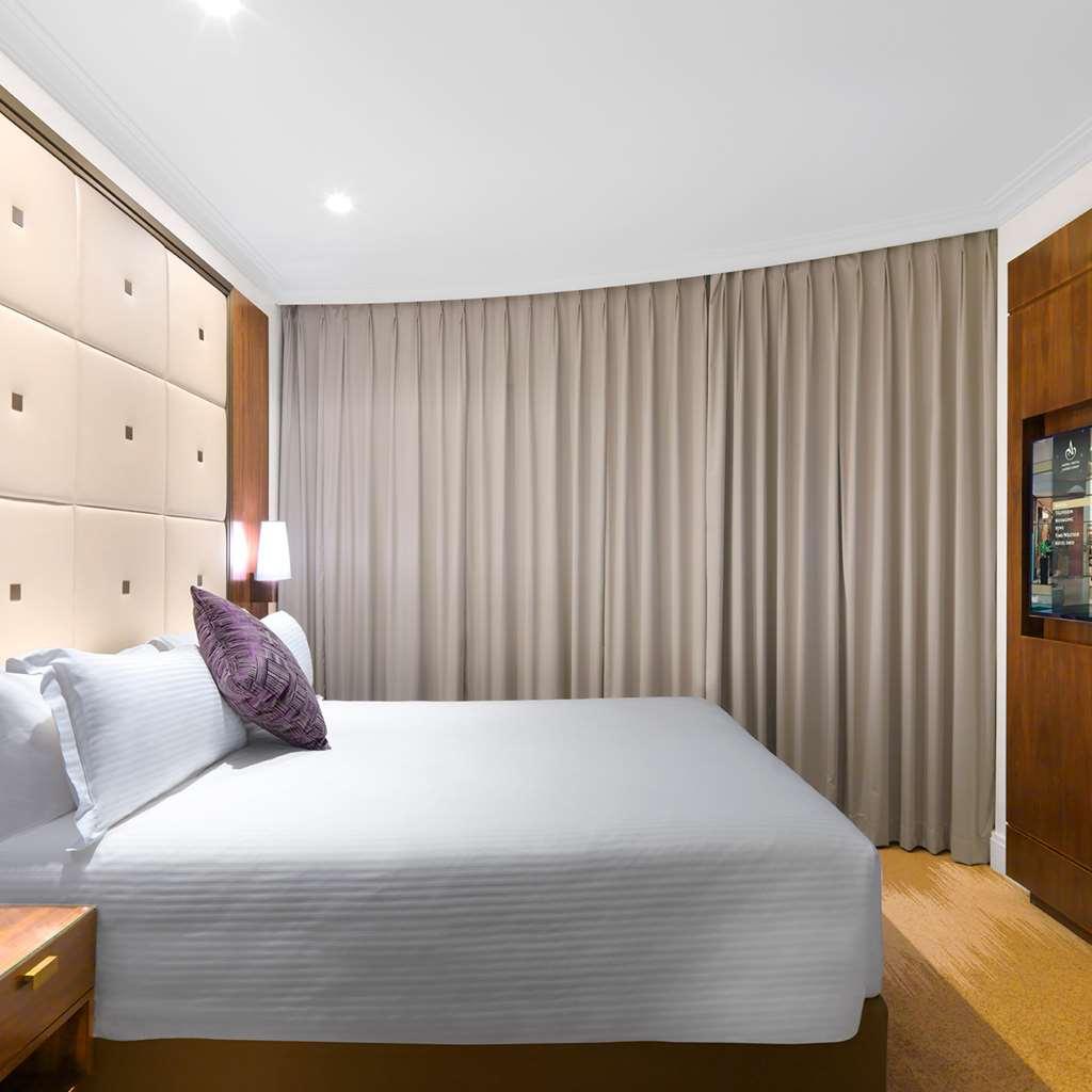 Amora Hotel Jamison Сідней Номер фото The photo shows a modern hotel room with a neatly made bed featuring light-colored bedding and a decorative pillow. The headboard behind the bed has a soft, upholstered design with square patterns. To the right, there is a wooden cabinet, and on the 
