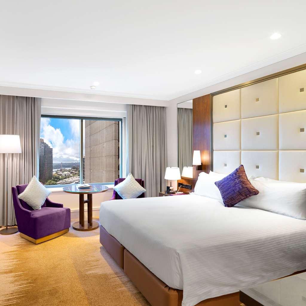 Amora Hotel Jamison Сідней Номер фото The photo shows a modern hotel room featuring a king-sized bed with white linens and a decorative purple pillow. To the side, there are two stylish armchairs in a deep purple color, accompanied by a small round table. The room has large windows cover