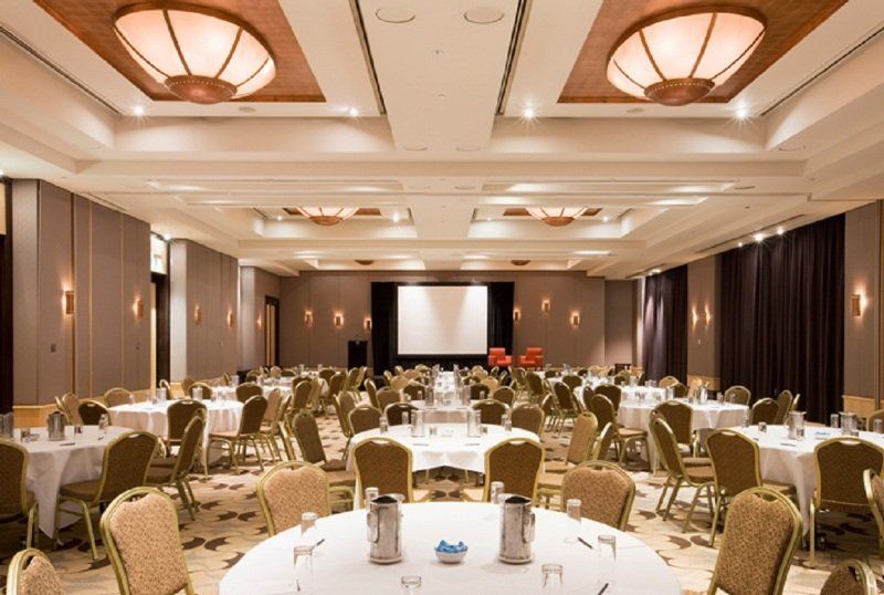 Amora Hotel Jamison Сідней Екстер'єр фото The photo shows a large banquet or conference room. There are several round tables arranged throughout the space, each covered with white tablecloths. The tables are set with water pitchers and glasses. In the center of the room, there is a projectio