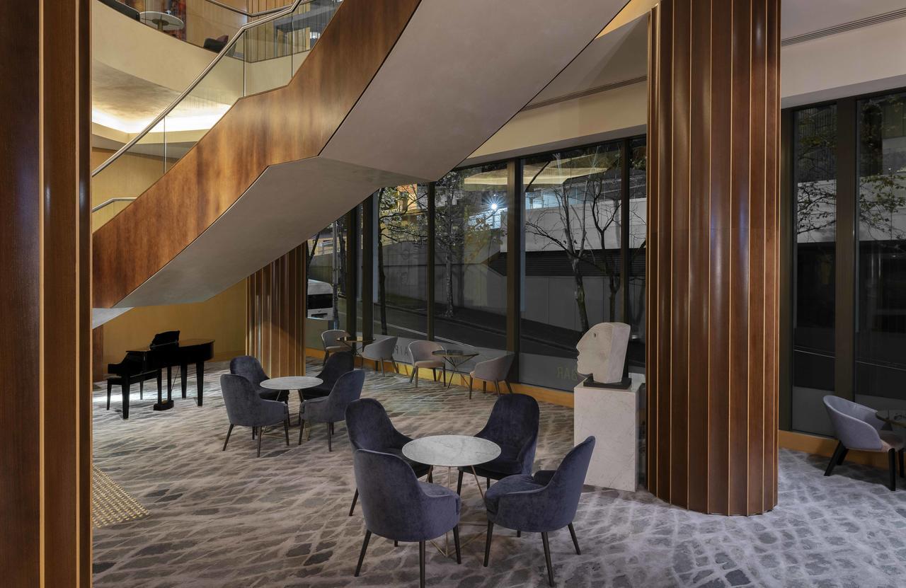 Amora Hotel Jamison Сідней Екстер'єр фото The photo depicts a modern interior space featuring a stylish lounge area. There are several comfortable chairs and small tables arranged on a patterned carpet. A large staircase with a sleek design is visible, leading to an upper level. The area is 