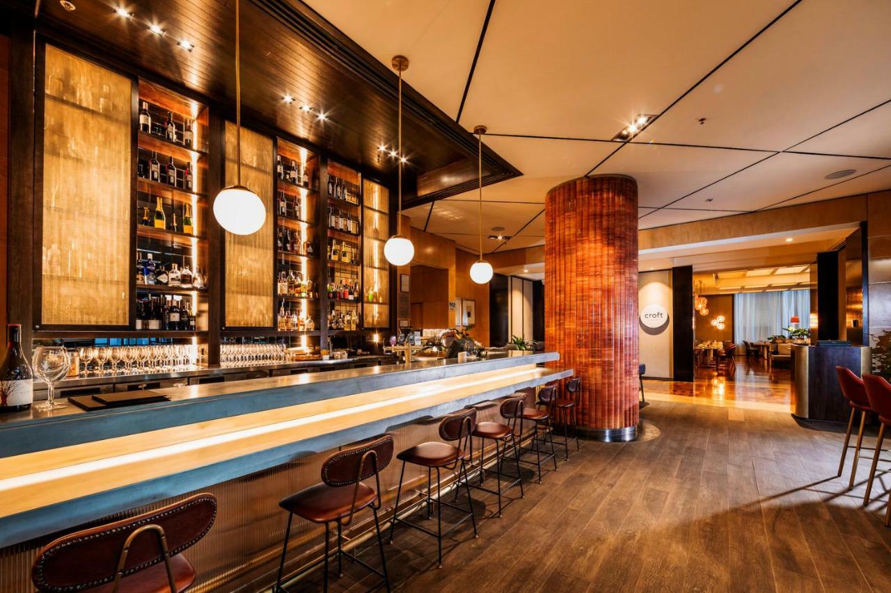 Amora Hotel Jamison Сідней Екстер'єр фото The photo shows the interior of a stylish bar. There is a long bar counter with a smooth surface, and several high-backed chairs lined up along it. Behind the bar, there is a wall of shelves filled with various types of bottles, likely containing spi