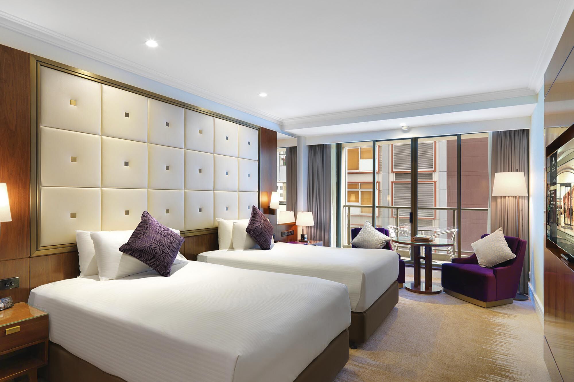 Amora Hotel Jamison Сідней Екстер'єр фото The photo depicts a modern hotel room featuring two neatly made beds with white linens and purple accent pillows. The walls are partially covered with a padded paneling, creating an elegant backdrop. There is a large window with sheer curtains allowi