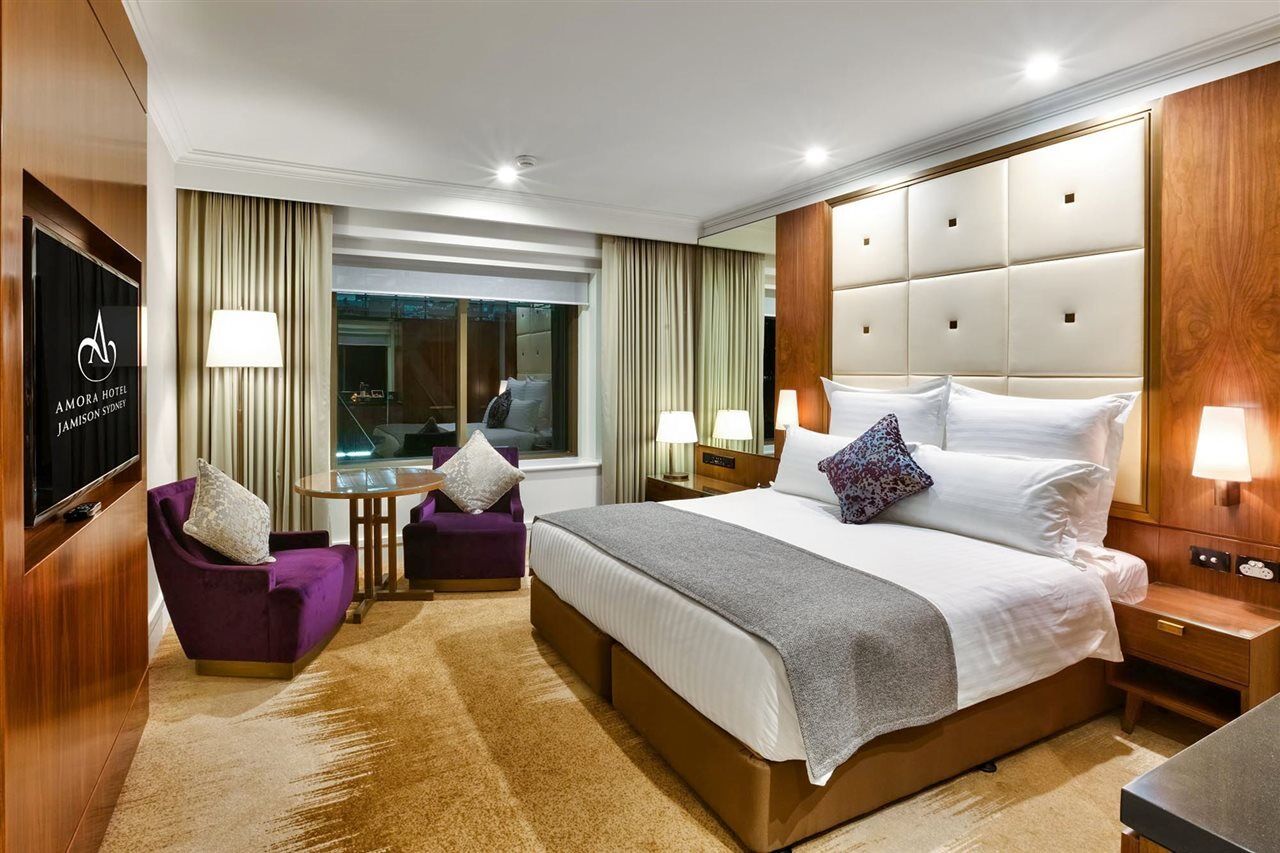 Amora Hotel Jamison Сідней Екстер'єр фото The photo shows a modern hotel room with a clean and stylish design. It features a large bed with white linens and a gray throw blanket. There are decorative pillows on the bed. To one side, there is a small seating area with two purple chairs and a 